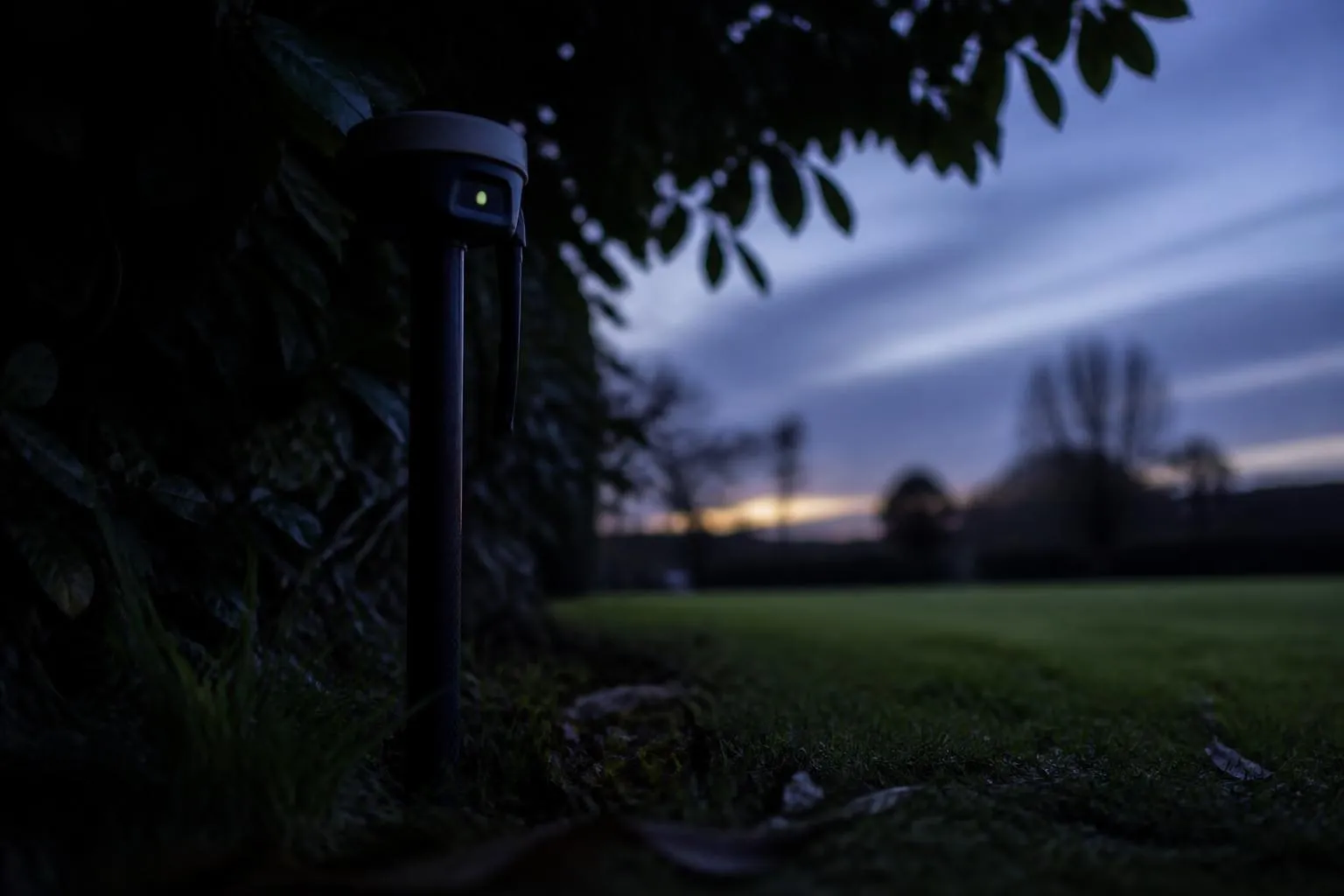 Mât d'antenne RTK planté au bord d'une pelouse au crépuscule, sous le feuillage refermé d'un grand arbre